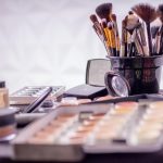 A collection of makeup products neatly arranged on a table, accompanied by a cup filled with makeup brushes. Caption: Explore the world of cosmetics through this image showcasing a variety of makeup products meticulously organized on a table. A cup filled with makeup brushes adds to the scene, hinting at the artistry and tools used in the application of makeup.